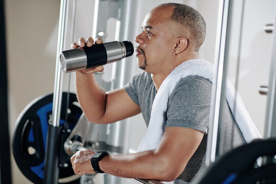 Mature man resting between sets, leaning on barbell and drinking vitamin water from thermos