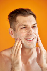 Obraz premium good-looking caucasian man cares for his skin, moisturizes it with professional cosmetics product, isolated orange background