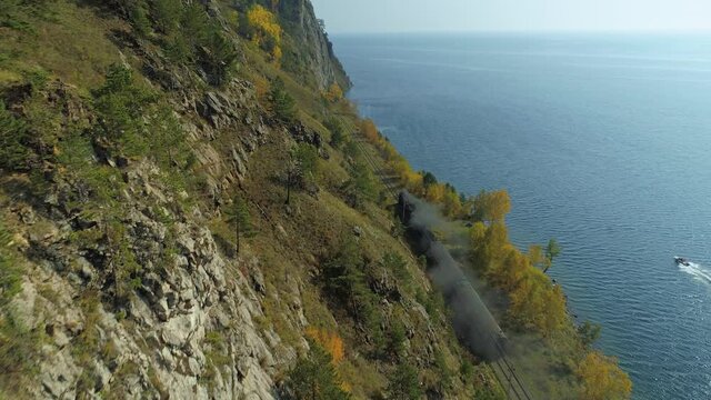 Old steam locomotive train rides along shore blue Lake Baikal Russia Circum-Baikal Trans-Siberian Railway. Epic autumn rocky natural landscape. Sunny. Open space horizon. Aerial forward flight film