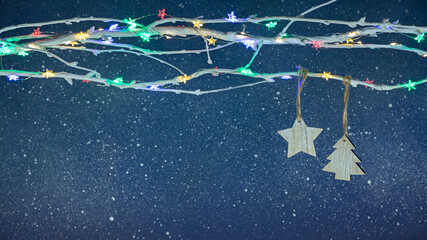 wooden christmas decorations hanging on bare tree branch decorated with colorful christmas lights against dark blue festive background