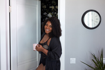 attractive plump black woman enter room holding cup of coffee, enjoy morning. in lingerie