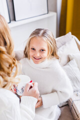 Mother and daughter in light sweaters are sitting on the sofa, the room is decorated for the new year and Christmas, hugs, conversations, smiles, kisses, waiting for a gift and magic