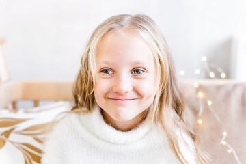 Baby girl in light sweater sitting on sofa, room decorated for new year and Christmas, waiting for gift and magic