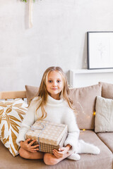 Baby girl in light sweater sitting on sofa, room decorated for new year and Christmas, waiting for gift and magic
