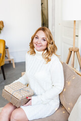 A woman with blonde hair in a white sweater sits on the sofa and holds a gift for the new year or Christmas, bright interior