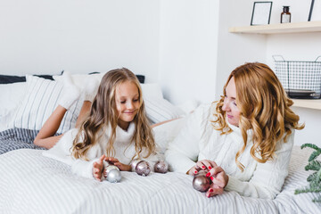 Mom and daughter blonde hair and white sweaters lie on the bed, light up, say toys for Christmas and new year tree, Christmas tree decoration