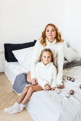 Mom and daughter blonde hair and white sweaters lie on the bed, light up, say toys for Christmas and new year tree, Christmas tree decoration