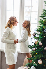 Girl child and mother are standing near the window, hugging, waiting for guests or Santa Claus for new year, Christmas, the room is decorated with a Christmas tree and balloons