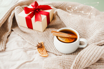 Obraz na płótnie Canvas cozy Christmas hygge style card. porcelain tea cup with orange slice, cinnamon sticks, anise star on beige crumpled blanket, gift with red ribbon and sparkling garland on background, selective focus