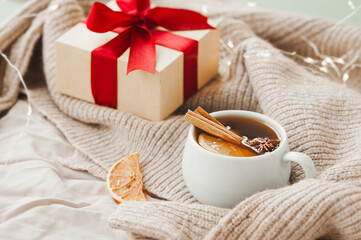 Obraz na płótnie Canvas cozy Christmas hygge style card. porcelain tea cup with orange slice, cinnamon sticks, anise star on beige crumpled blanket, gift with red ribbon and sparkling garland on background, selective focus