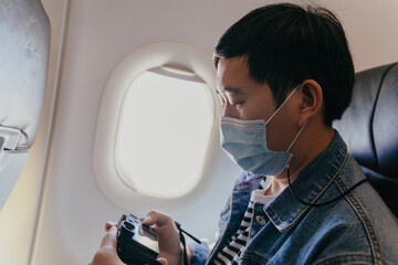 Fototapeta na wymiar 20s young adult Asian male tourist wearing a face mask inside aircraft cabin air and taking photos with a camera. Tourism during Covid-19 or Coronavirus pandemic. New normal travel concept