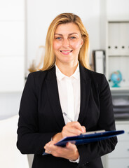 Young adult female assistant standing with clipboard and making notes in modern office