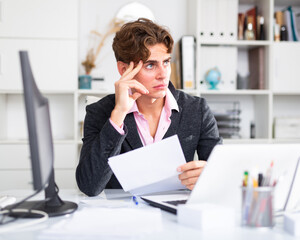 Attractive thoughtful young man working with documents and laptop in office