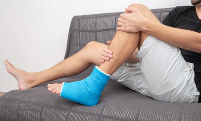Fototapeta premium Man with broken leg in blue splint for treatment of injuries from ankle sprain on couch at home.