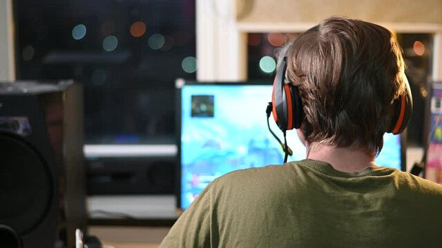 The guy plays a computer game online with excitement at home in the room. Back view