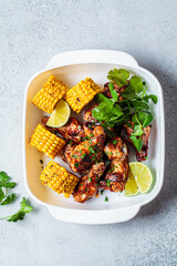 Barbecue chicken wings served with grilled corn and greens in white dish, gray background.