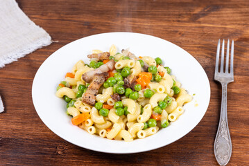 Traditional italian chifferini pasta with peas, carrots and toasted pancetta on a plate on a rustic wooden table, background