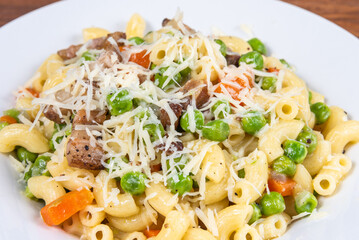 Traditional italian chifferini pasta with peas, carrots and toasted pancetta, sprinkled with grated cheese on a plate on a rustic wooden table, background