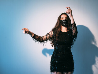 Pandemic party. Night club look. Fashion trend 2021. Brunette woman in black handmade face mask matching cocktail dress enjoying dancing alone on blue light empty space background.
