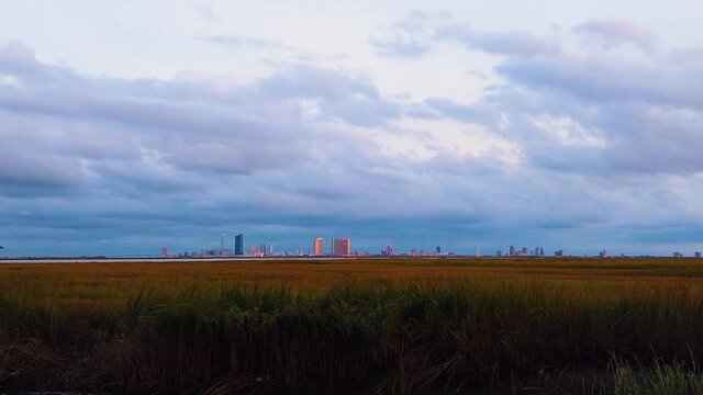 HD MOTIONLAPSE Zoom out Atlantic City skyline in distance over waterway with mostly cloudy sky day to night with clouds turning pink, purple and blue