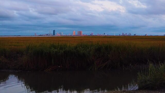 HD MOTIONLAPSE Straight tilt up and hold Atlantic City skyline in distance over waterway with mostly cloudy sky day to night with clouds turning pink, purple and blue