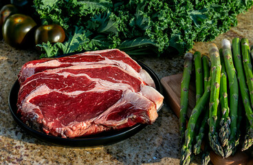 Raw meat, beef steak on black plate with kale, asparagus, broccolini, and tomatoes in the background.