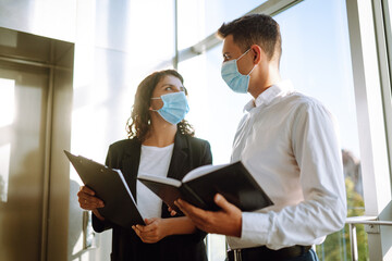 Two coworkers in protective face masks discussing project, sharing ideas, having pleasant conversation near elevator. Work in office during quarantine. Covid-19.