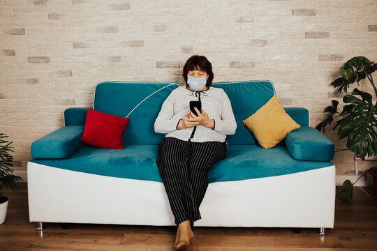 Senior Woman Wearing Protective Medical Mask Sitting On The Sofa In The Living Room At Home. Stay Home For Protection From Virus.