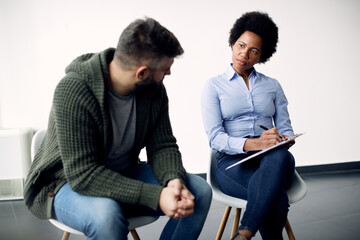 Black female psychotherapist taking notes while talking to a man during counselling.