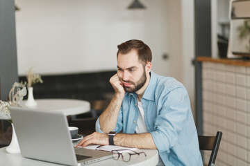 Dissatisfied boring young man sitting alone at table in coffee shop cafe restaurant indoor working...