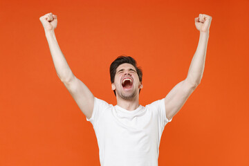 Fototapeta premium Overjoyed screaming young man in basic casual white blank empty t-shirt standing clenching fists doing winner gesture looking camera isolated on bright orange colour wall background, studio portrait.
