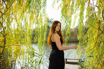 A blonde woman in a black long dress on a wooden pier on the river bank. Beautiful landscapes, green leaves and trees on the shore