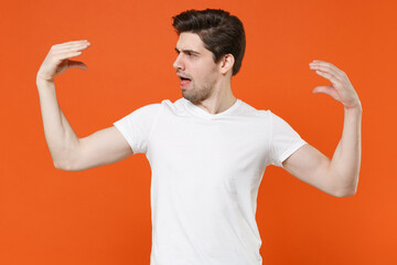 Shocked perplexed puzzled young man 20s wearing basic casual white blank empty t-shirt standing showing blah blah gesture ja jaja hands isolated on bright orange colour background, studio portrait. © ViDi Studio