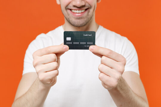 Cropped image of smiling cheerful handsome young man 20s wearing basic casual empty blank white t-shirt holding in hands credit bank card isolated on bright orange colour background studio portrait.