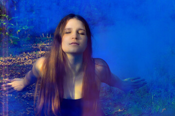 Fototapeta na wymiar Red-haired woman sitting in a black long dress in a puff of blue smoke in the forest. Mysticism