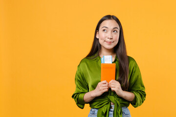 Pensive pretty young traveler tourist asian woman in green shirt hold passport tickets looking up isolated on yellow background. Passenger traveling on weekends getaway. Air flight journey concept.