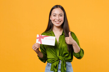 Smiling funny young brunette asian woman wearing basic green shirt standing hold in hand gift certificate showing thumb up looking camera isolated on bright yellow colour background, studio portrait.
