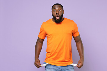 Shocked worried young african american man 20s wearing basic casual blank orange t-shirt standing has no money showing empty pockets isolated on pastel violet colour background studio portrait.