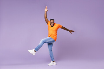 Fototapeta na wymiar Full length of cheerful funny young african american man 20s wearing basic casual orange t-shirt dancing rising leg spreading hands looking camera isolated on pastel violet background studio portrait.