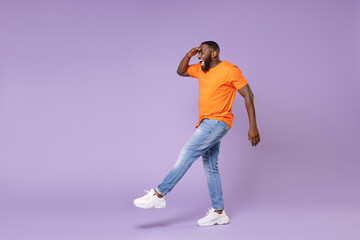 Fototapeta na wymiar Full length of excited funny young african american man 20s wearing basic casual orange t-shirt holding hand at forehead looking far away distance isolated on pastel violet background studio portrait.