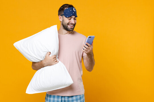 Smiling young man in pajamas home wear sleep mask hold pillow using mobile cell phone typing sms message while resting at home isolated on yellow background studio portrait. Relax good mood concept.