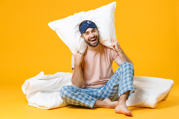 Full length of laughing young bearded man in pajamas home wear sleep mask sitting with pillow...