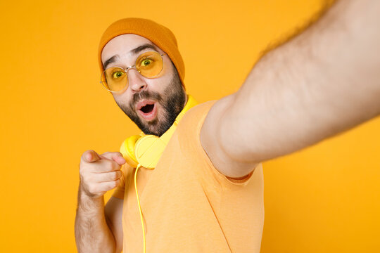 Close up of shocked young bearded man in basic casual t-shirt headphones eyeglasses hat doing selfie shot on mobile phone pointing index finger on camera isolated on yellow background studio portrait.