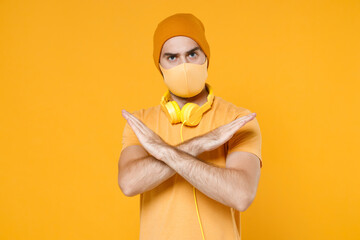 Worried young man in t-shirt hat headphones face mask to safe from coronavirus virus covid-19 during quarantine showing stop gesture with crossed hands isolated on yellow background studio portrait.