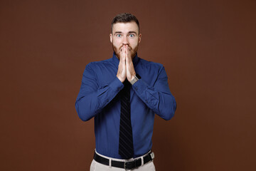 Shocked amazed bearded young business man wearing blue shirt tie covering mouth with hands looking camera isolated on brown background studio portrait. Achievement career wealth business concept.