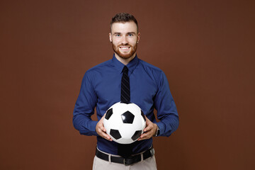 Smiling young business man football fan in blue shirt cheer up support favorite team with soccer ball looking camera isolated on brown background studio portrait. Achievement career wealth concept.