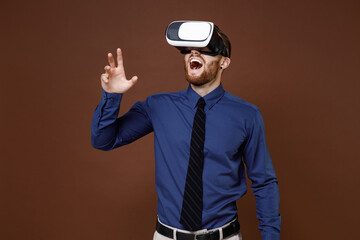 Excited young business man in blue shirt tie watching in vr headset gadget touch something like push click on button isolated on brown background studio. Achievement career wealth business concept.