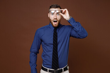 Shocked amazed bearded young business man wearing blue shirt tie eyeglasses keeping mouth open looking camera isolated on brown background studio portrait. Achievement career wealth business concept.