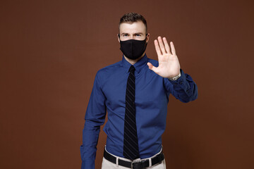 Displeased young business man in blue shirt face mask to safe from coronavirus virus covid-19 showing STOP gesture with palm isolated on brown background. Achievement career wealth business concept.