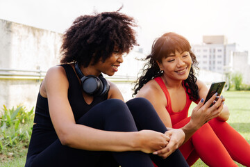 Diverse group of two female fitness friends talking and using a mobile phone in the park. Young women resting and smiling outdoors